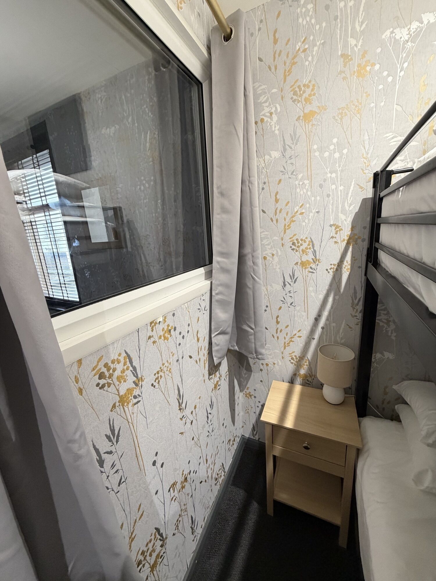 A bedroom with a window, a bedside table with a lamp, and floral wallpaper.