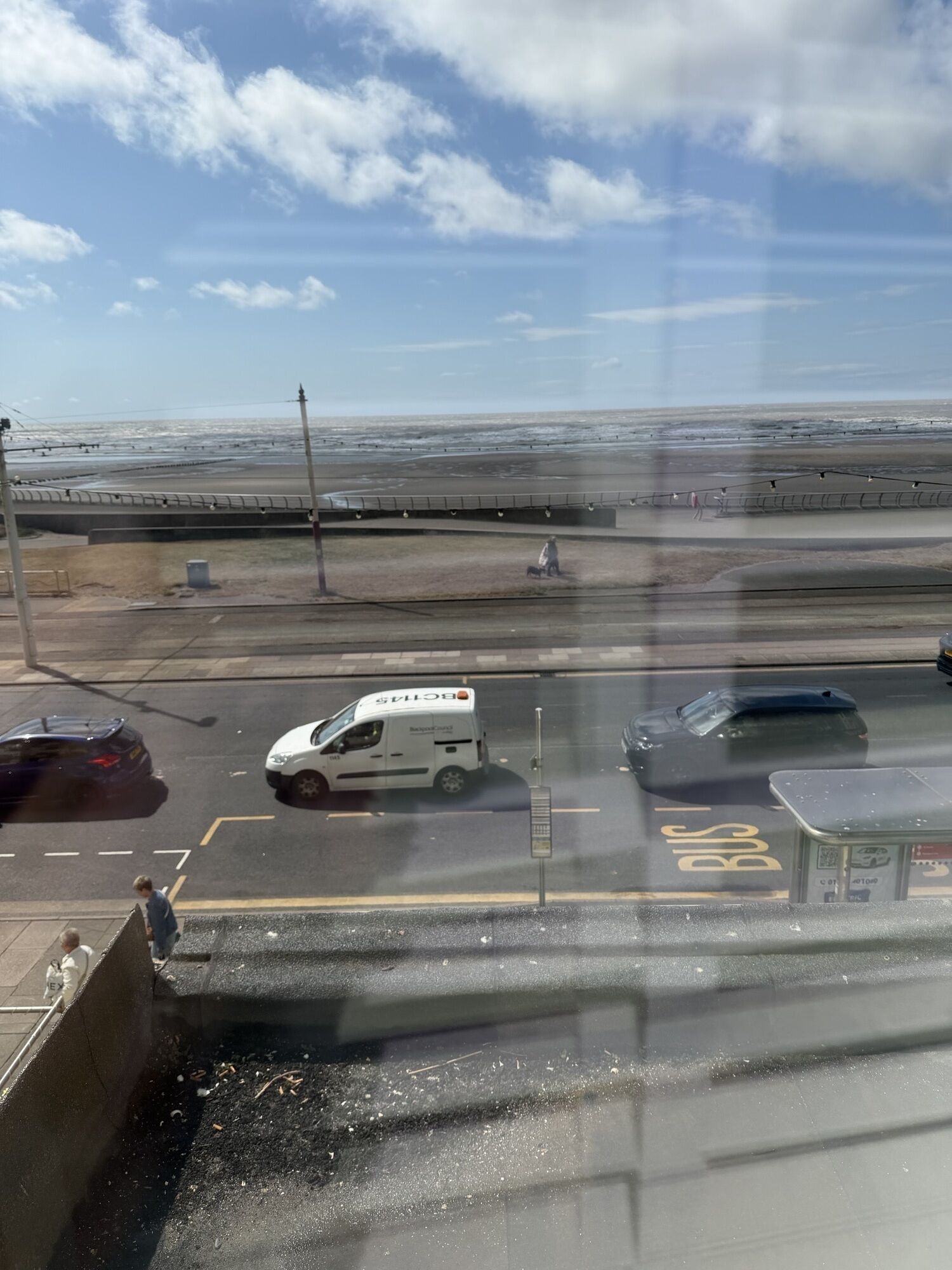 Un’area di parcheggio con un autobus e un furgone bianco, una strada con un attraversamento pedonale e vista sulla spiaggia e sul mare.