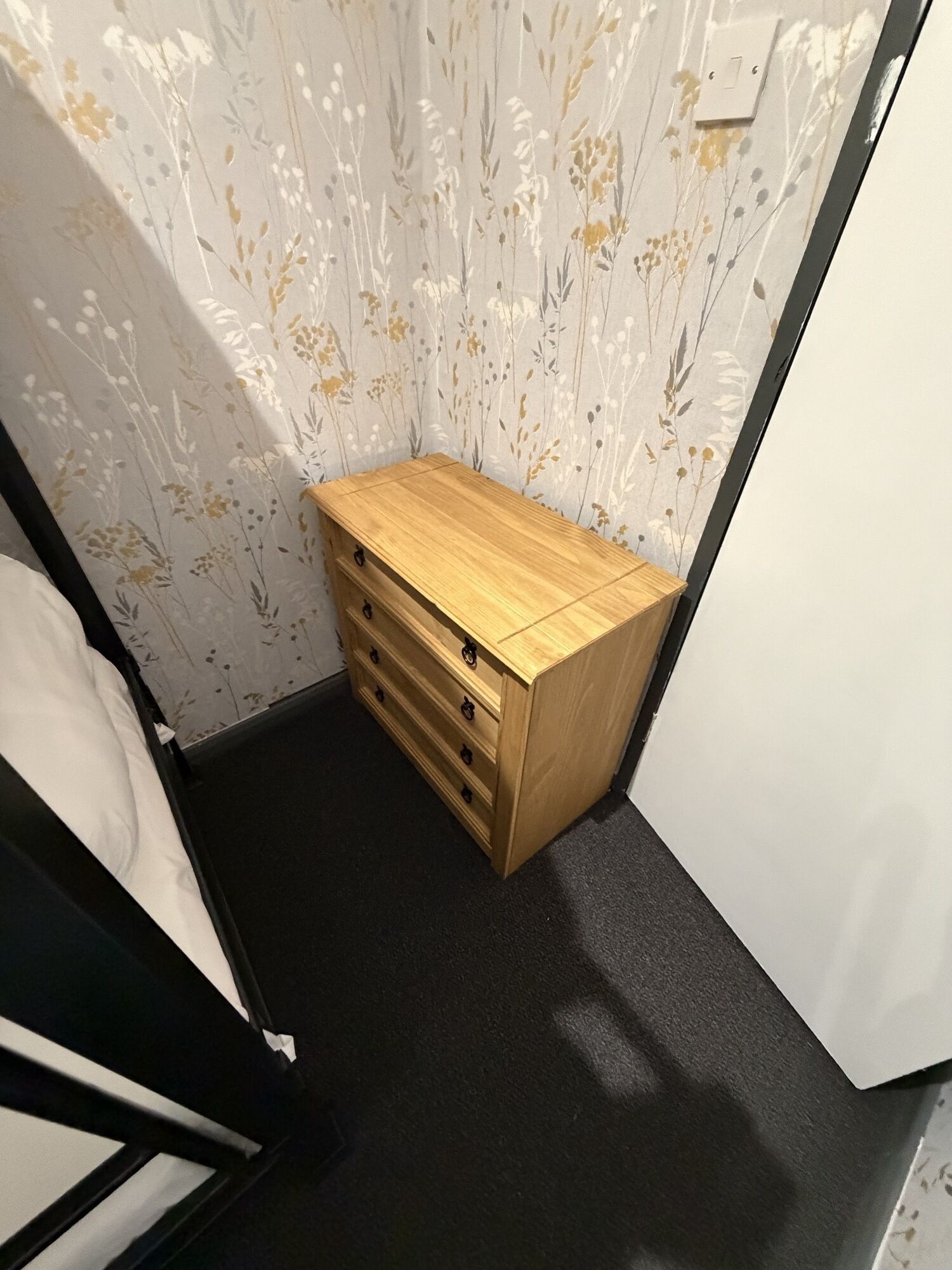 A corner bedroom with a wooden dresser, a bed, and floral wallpaper.