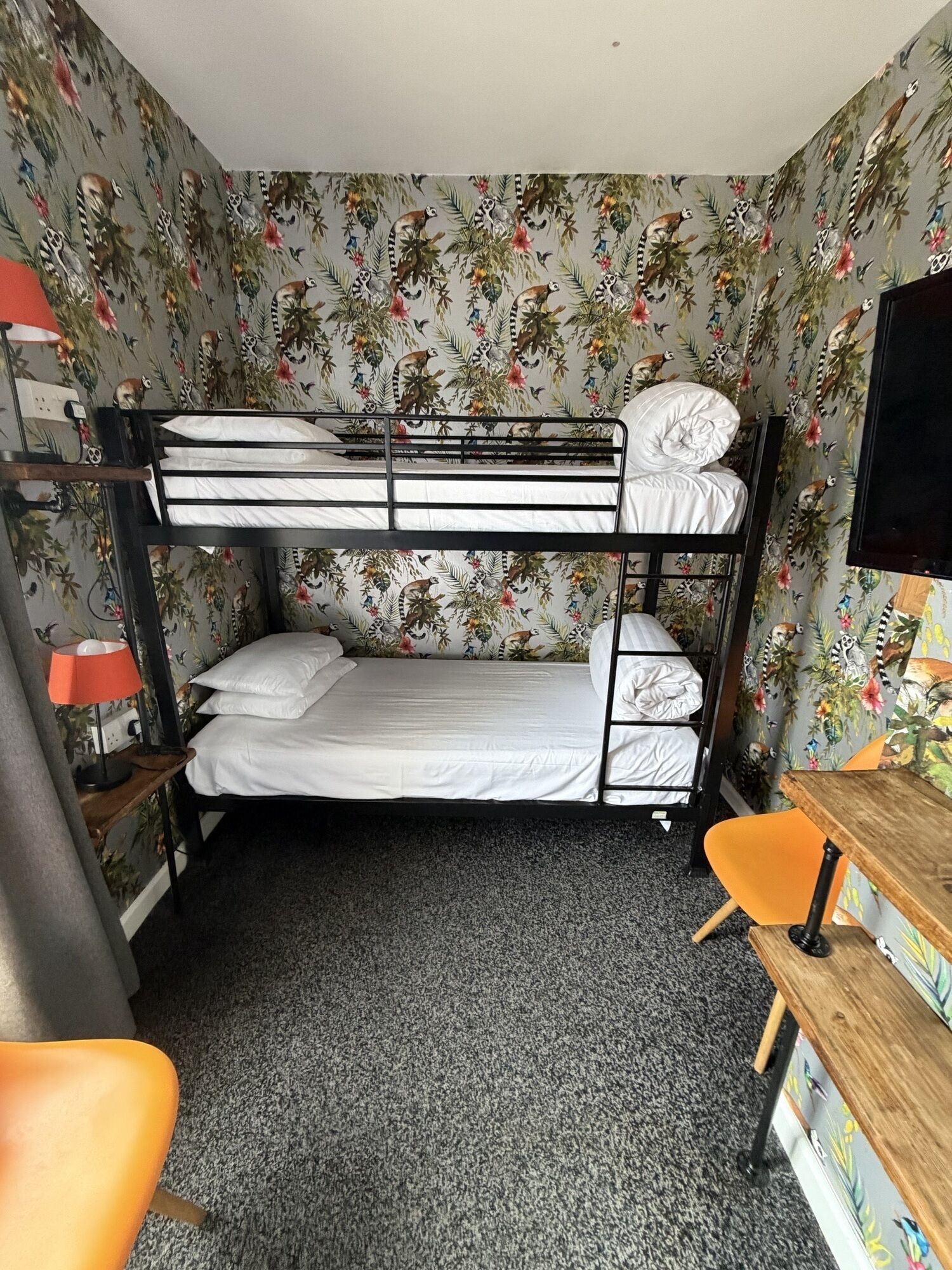 A bunk bed room with a floral wallpaper, a desk with a chair, and a television.