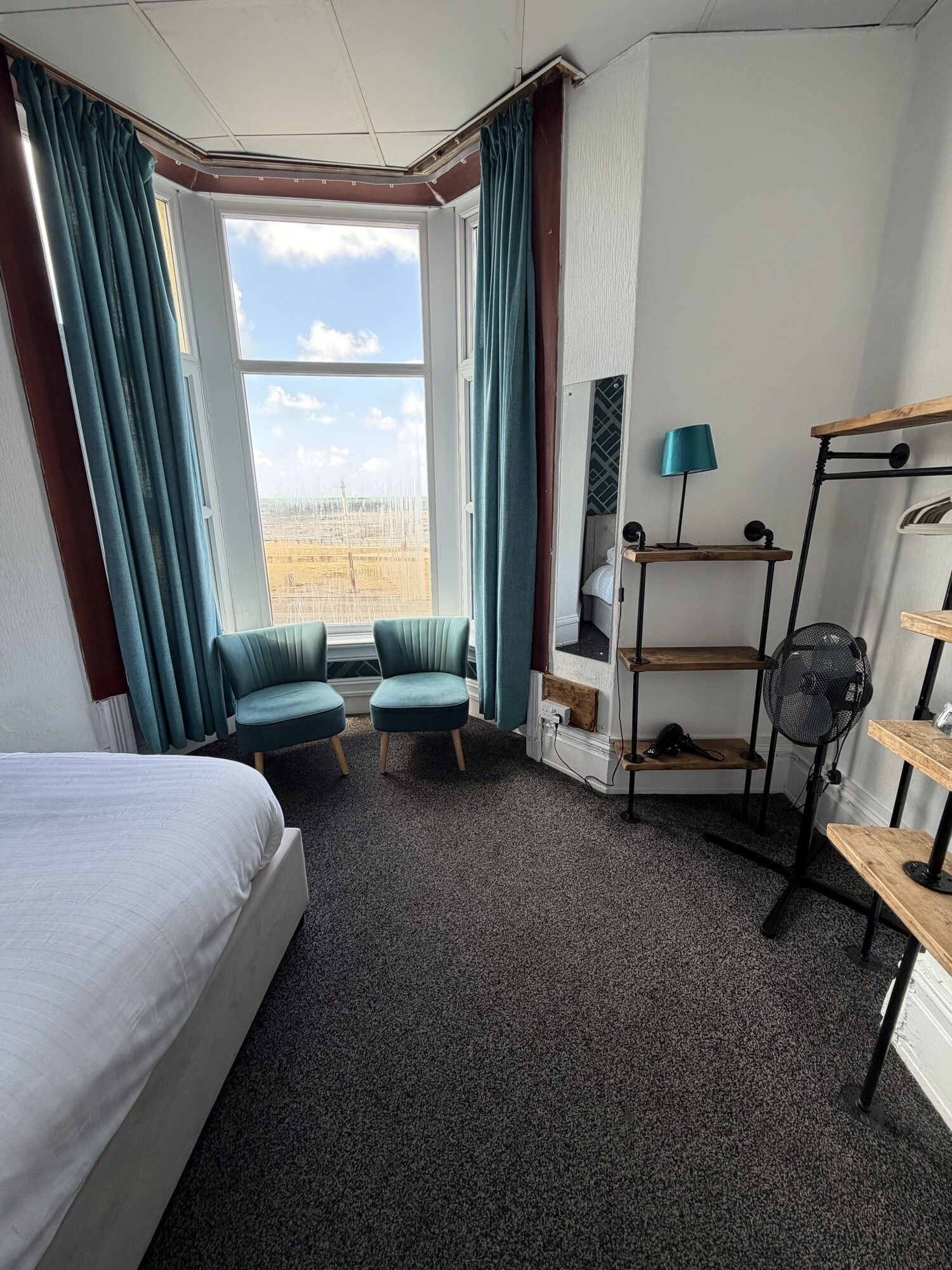 A hotel room with a bed, two armchairs, a standing fan, a wooden shelf, and a view of the cityscape through a large window.