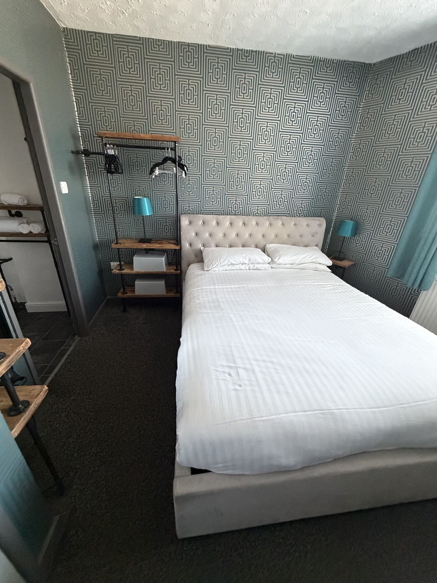 A hotel room with a large bed, a bedside table, a lamp, a wardrobe, and a patterned wall.