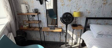 Habitación de hotel con cama, escritorio con espejo, una silla, ventilador de pie y una mesita de noche con lámpara.