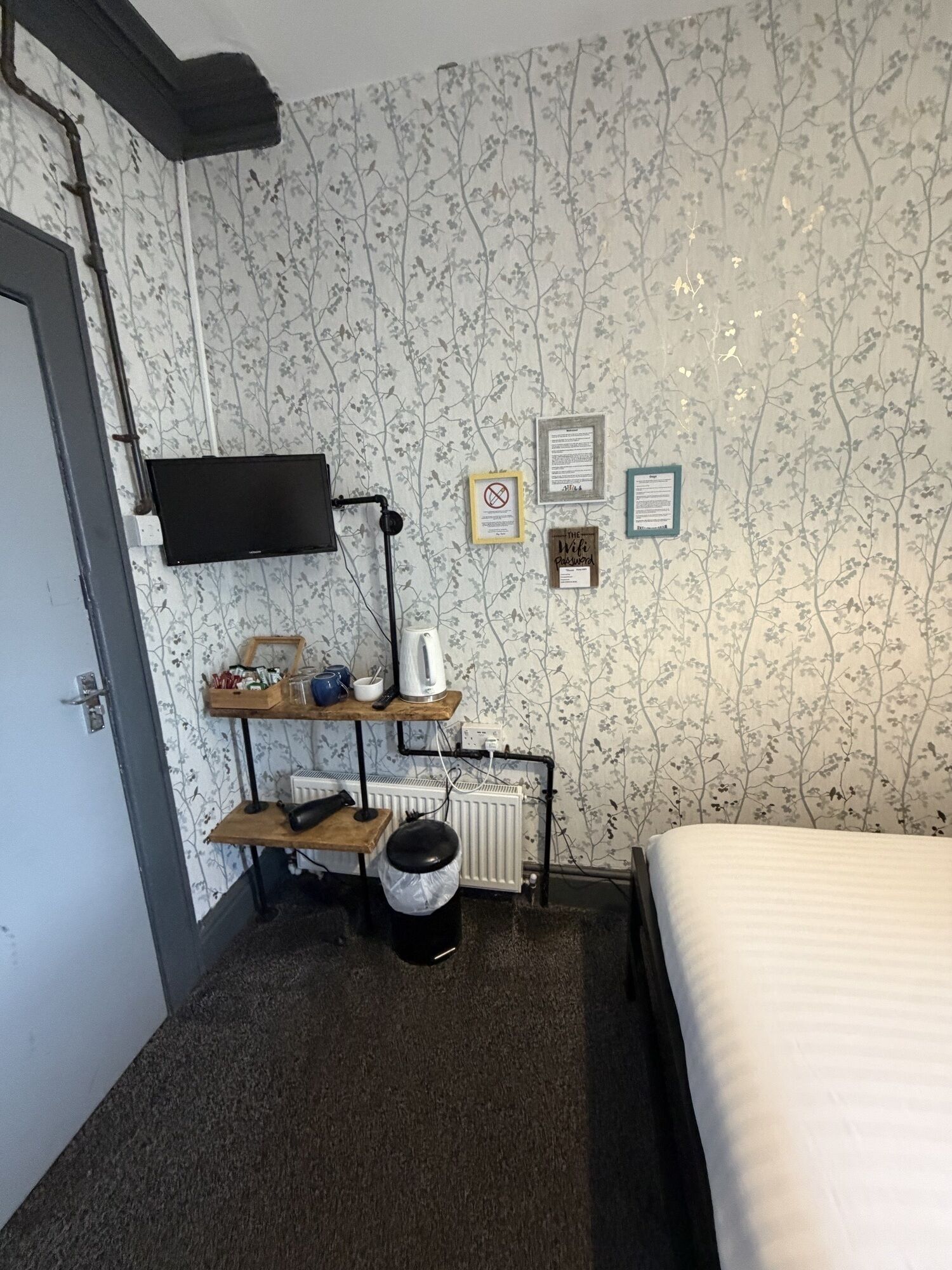 A small hotel room with a bed, a desk with a computer, a radiator, and a TV mounted on the wall.
