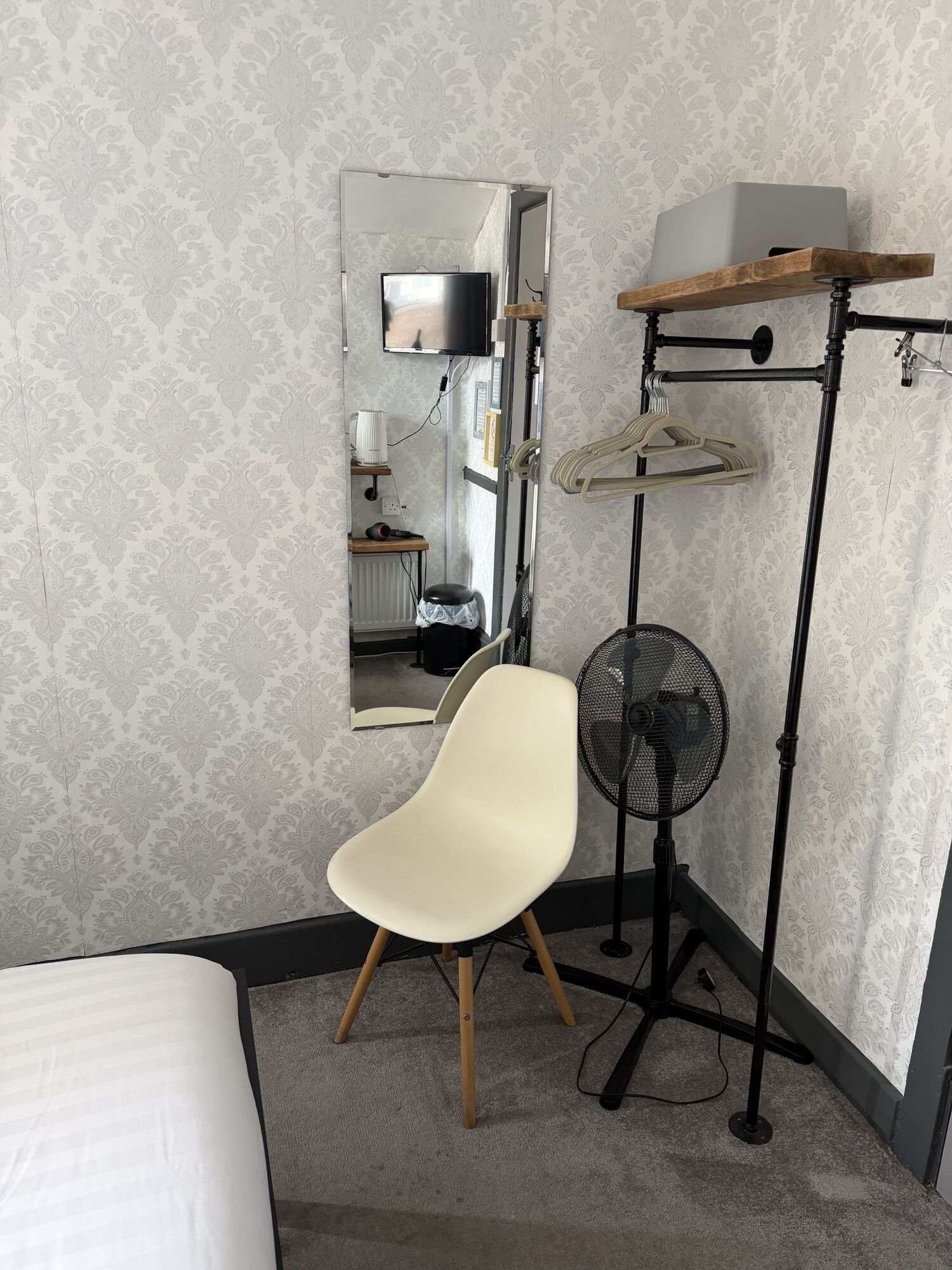 Una habitación con cama, una silla, un espejo, televisor, ventilador y una estantería con perchas.