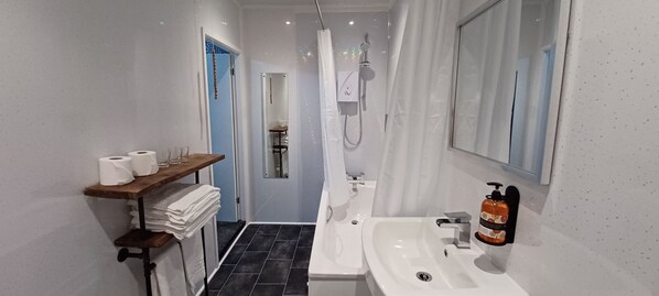 Ein modernes Badezimmer mit einem weißen Waschbecken, einer Dusche und einem Spiegel. Handtücher und Toilettenpapier liegen auf einem Regal.