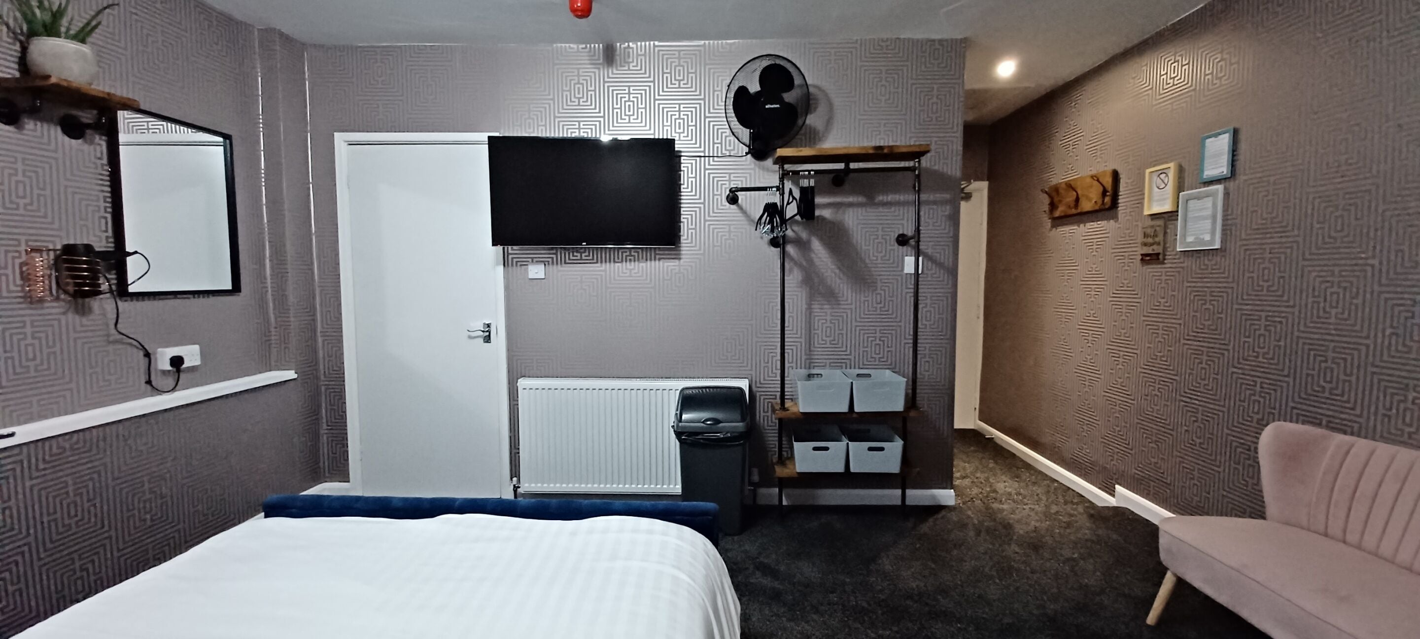 Una camera d'albergo con un letto, una TV fissata al muro, un termosifone, una scrivania con una sedia, uno scaffale con contenitori e una sedia rosa.