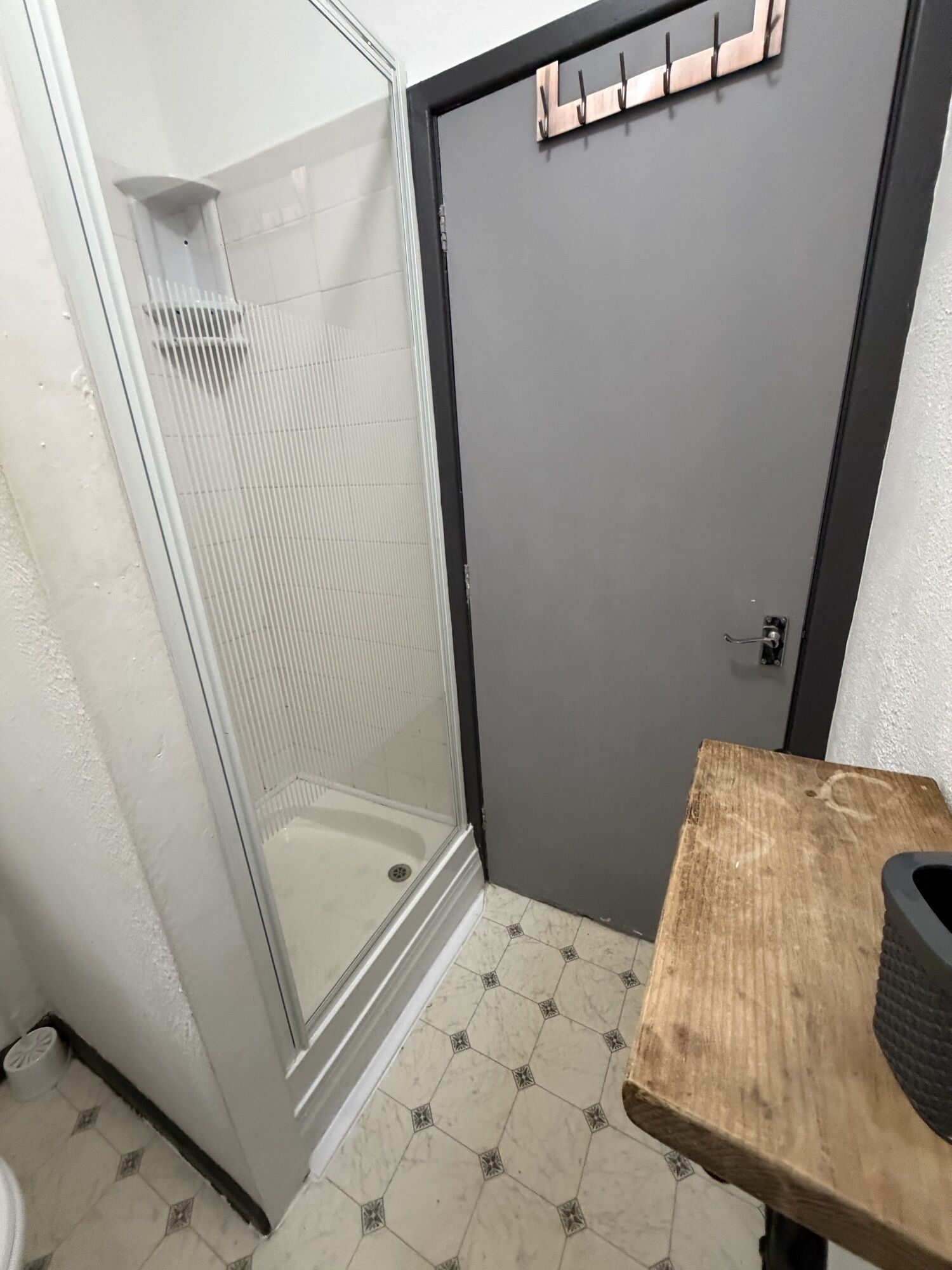 Un baño con cabina de ducha de vidrio, una puerta gris y una pequeña mesa de madera con un objeto negro encima.