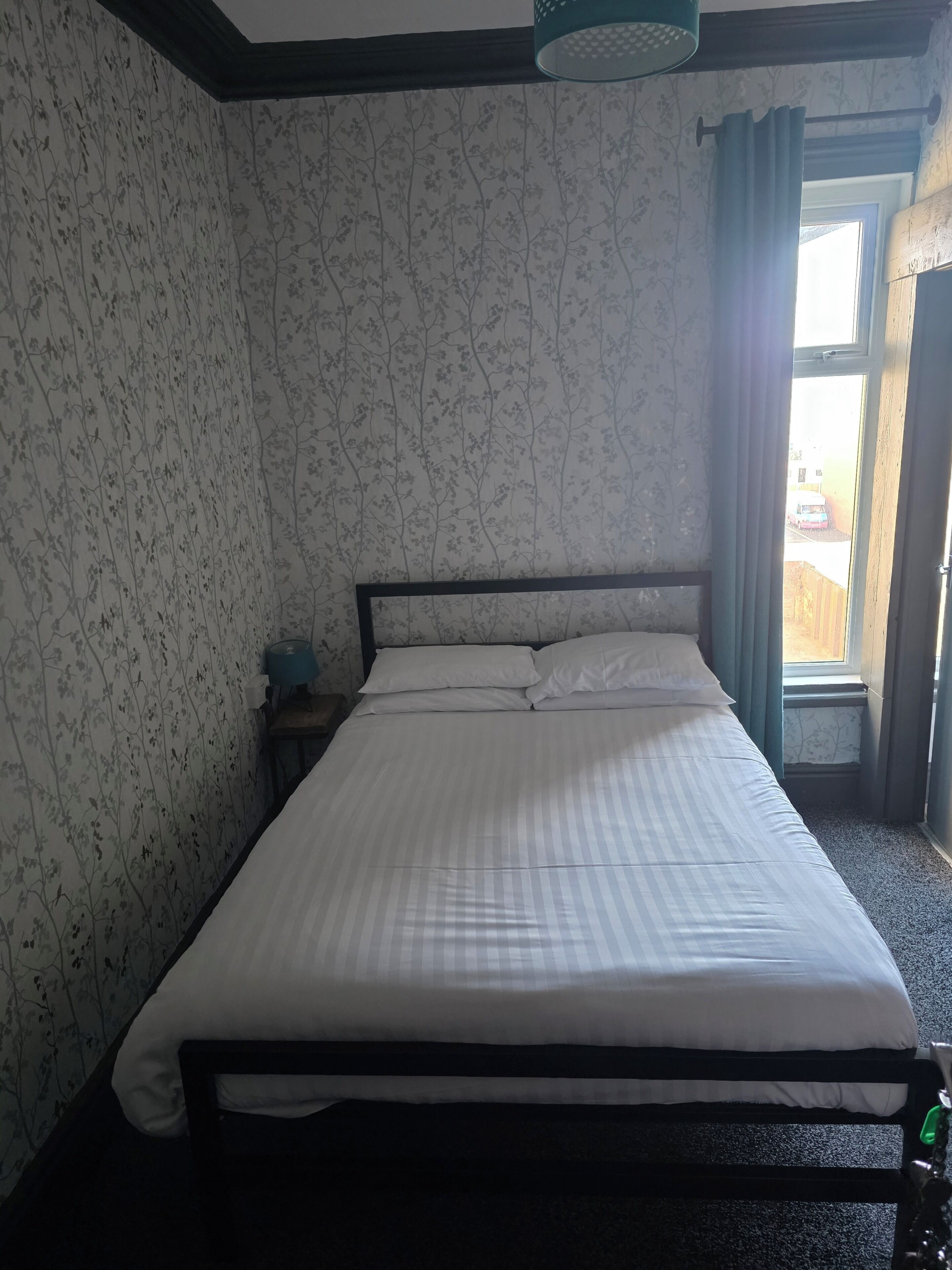 A bedroom with a bed, a nightstand, a window with curtains, and a view of the outside.