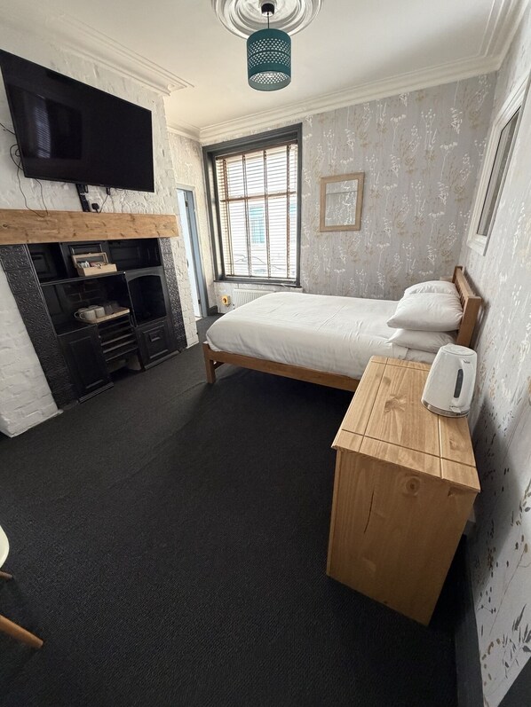 A bedroom with a bed, a wooden bench, a kettle, a TV, and a window with blinds.