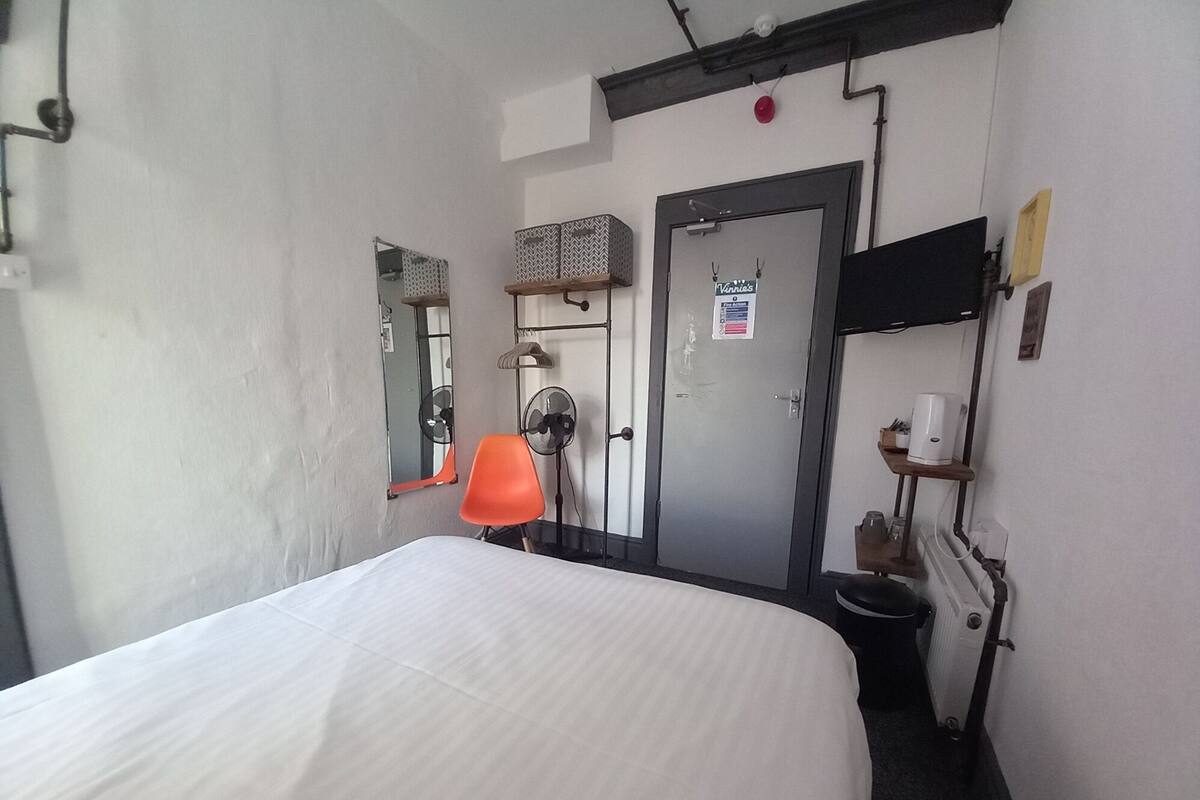 Habitació d'hotel amb un llit, una cadira, una porta, un mirall, un ventilador i un radiador.