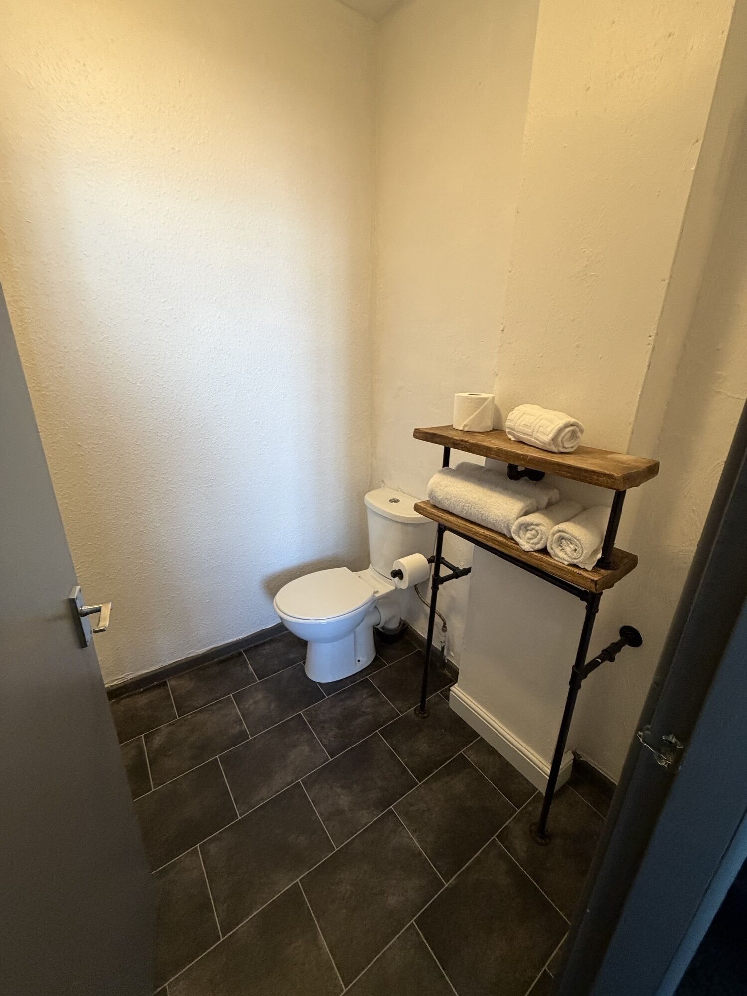A small bathroom with a toilet, a shelf with towels, and dark tiled flooring.