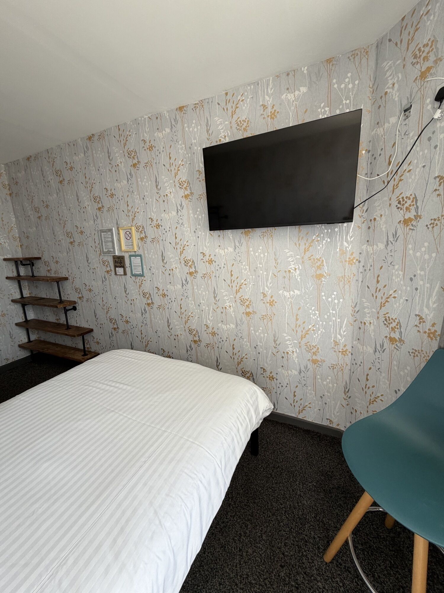 A bedroom with a bed, a desk, a chair, a TV mounted on the wall, and a patterned wallpaper.