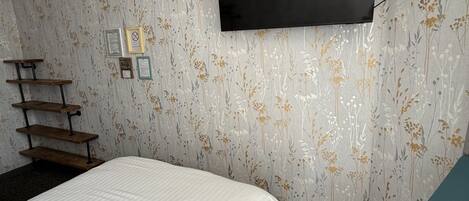 A bedroom with a bed, a desk, a chair, a TV mounted on the wall, and a patterned wallpaper.