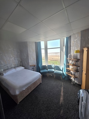A hotel room with a bed, two chairs, a small table, a shelf with items, a window with a view of the beach, and a ceiling with a grid pattern. - Vinnies Family Hotel (Blackpool)