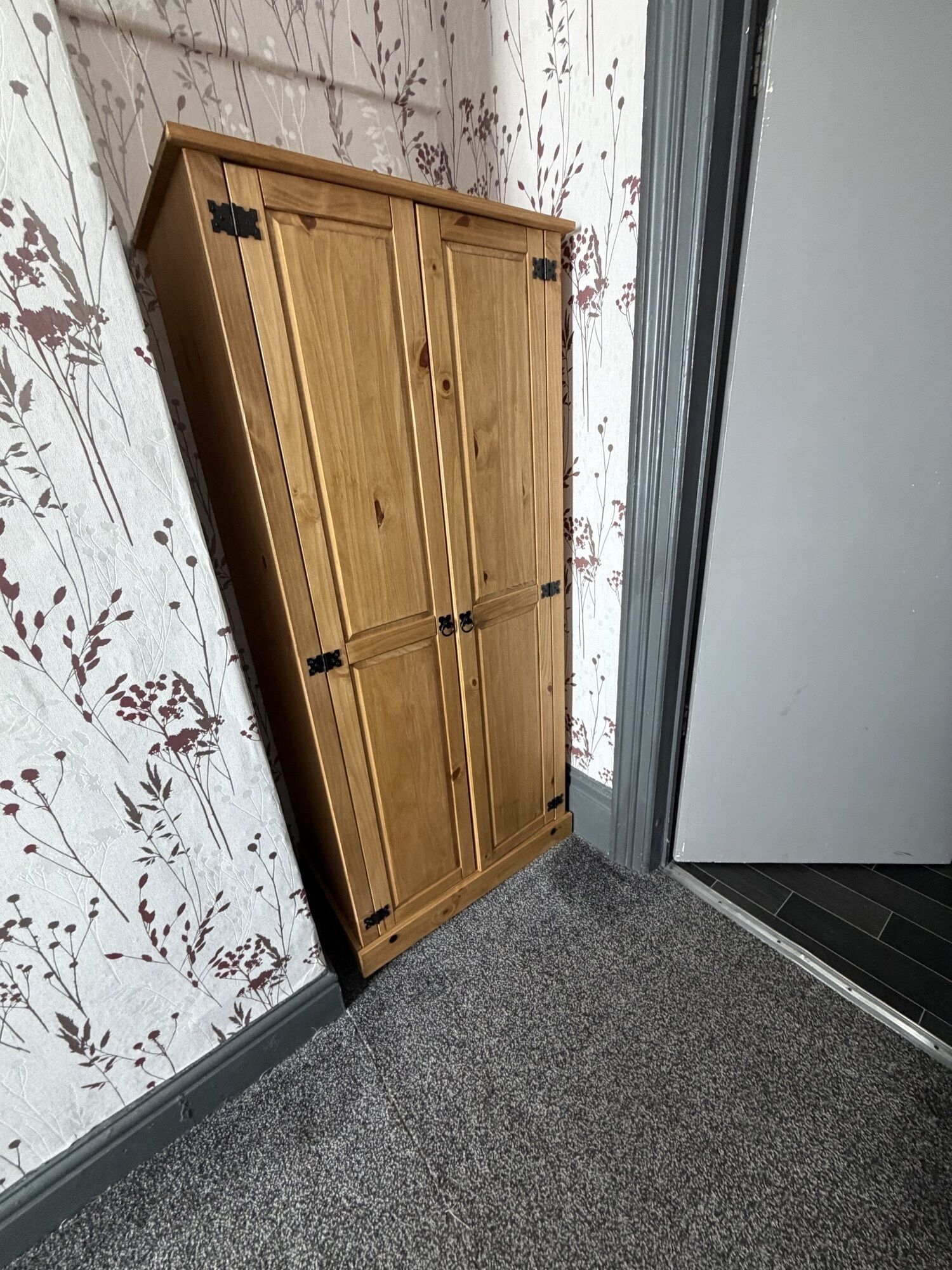 A corner with a wooden wardrobe against a floral wallpaper, a grey carpet, and a grey door.