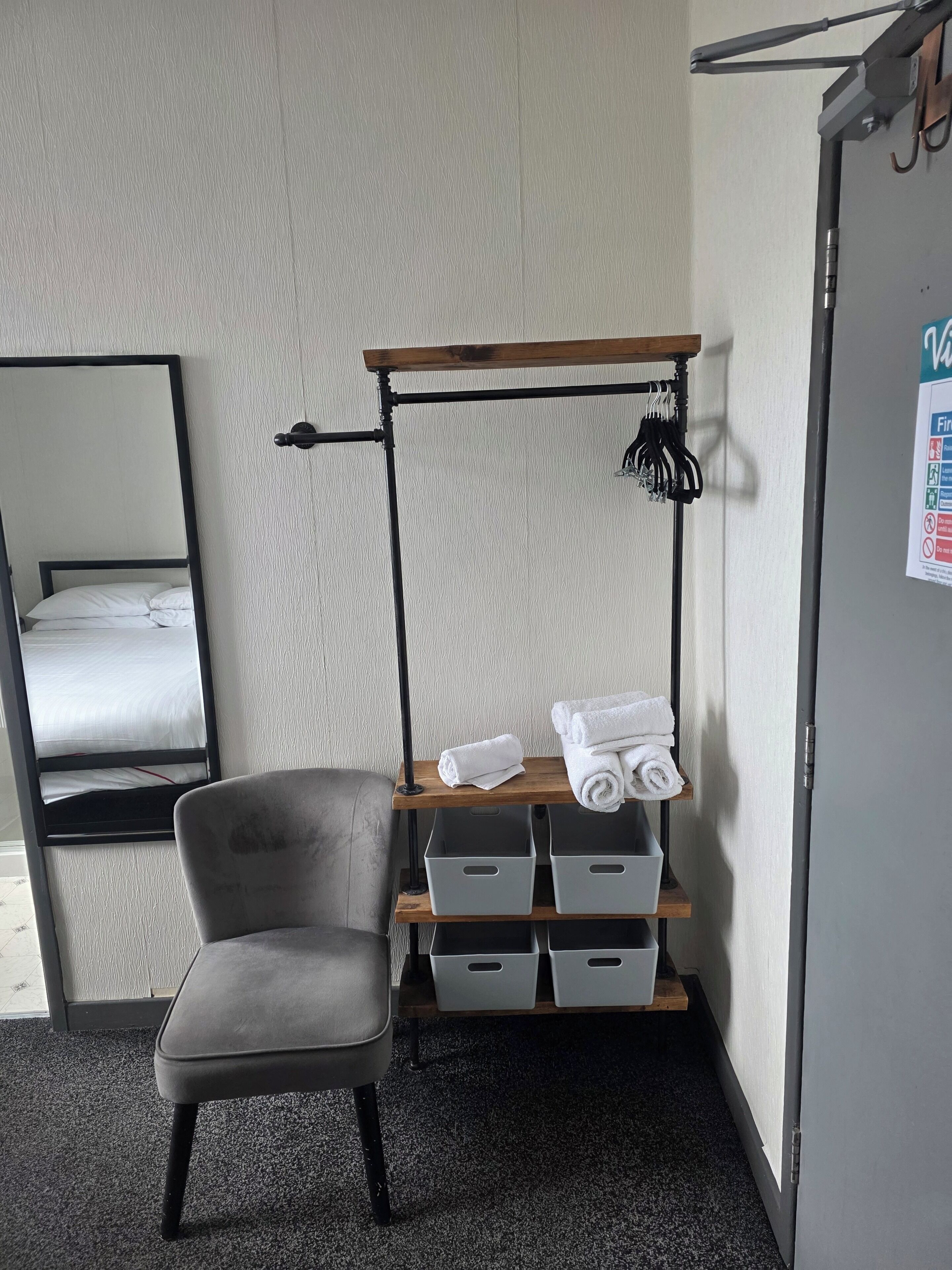 A hotel room with a bed, a chair, a shelf with towels, and storage bins.