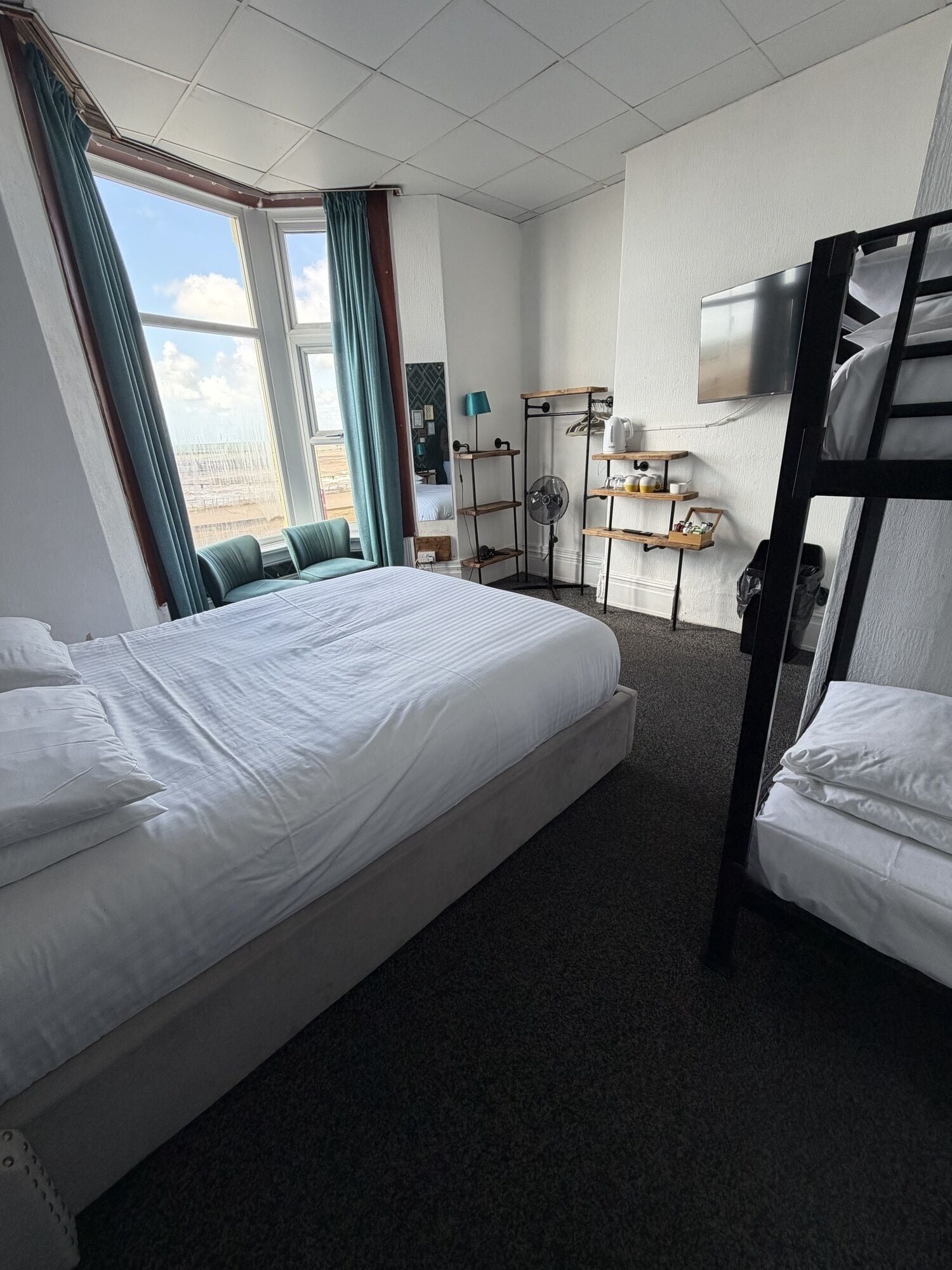 Habitación de hotel con una cama grande, una litera, un escritorio con lámpara y una ventana con cortinas.