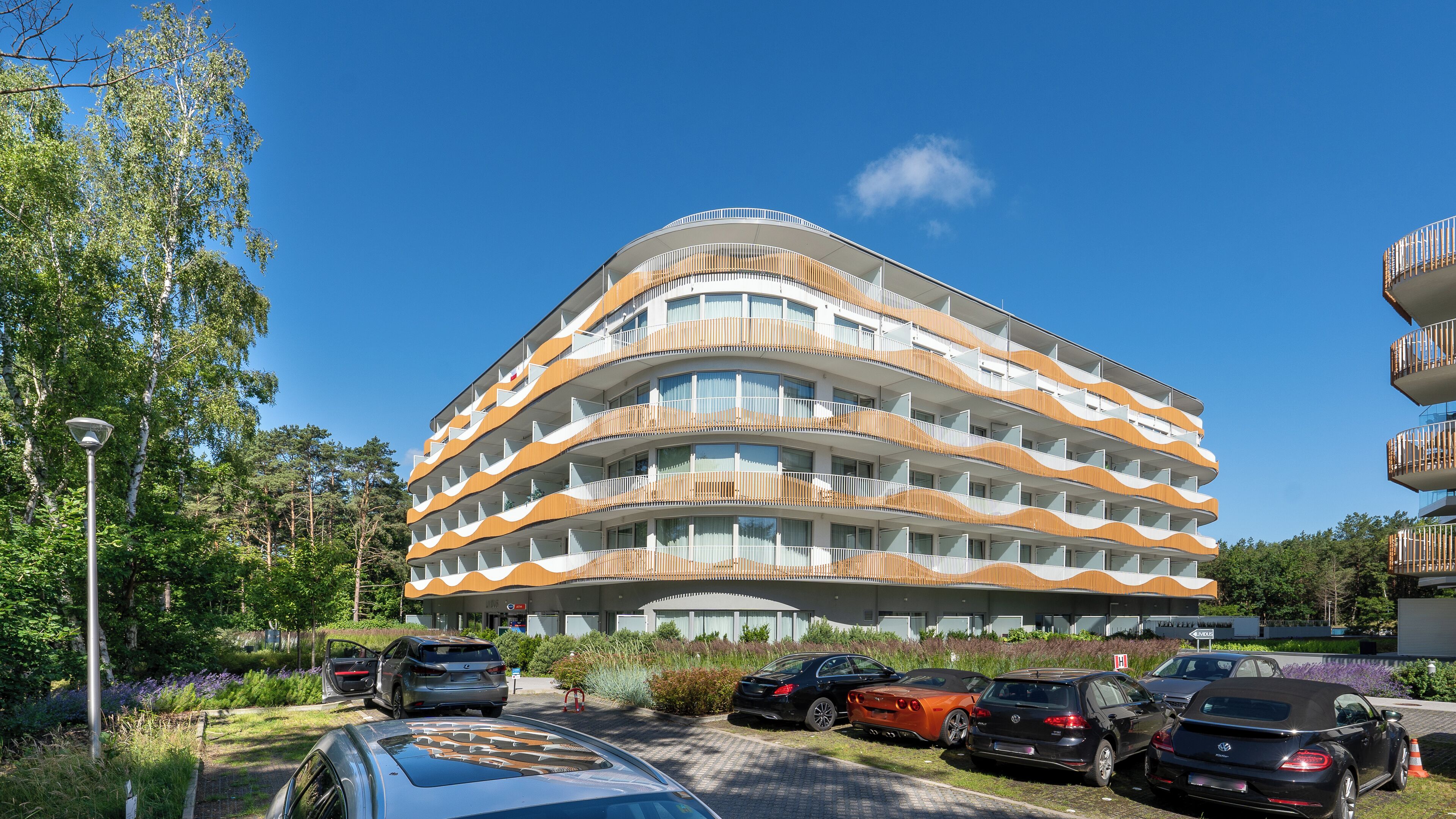 A multi-story building with a curved facade, balconies, and a parking area with cars.