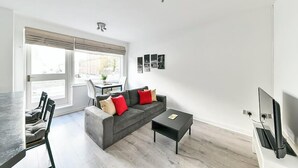 A modern living room with a grey sofa, a coffee table, and a TV. There is a dining area with a table and chairs. - The Elden - No.3 (London)
