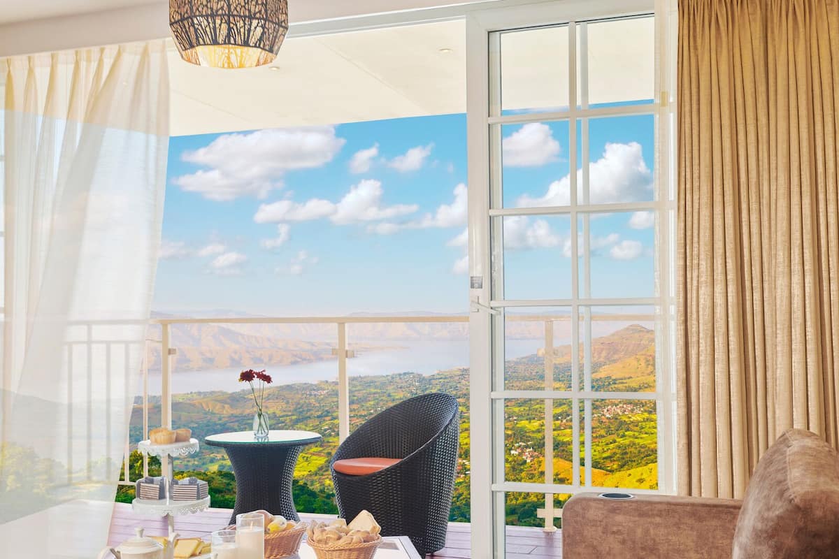 A balcony with a table set for tea, a wicker chair, and a sofa, overlooking a scenic landscape with mountains and a body of water.
