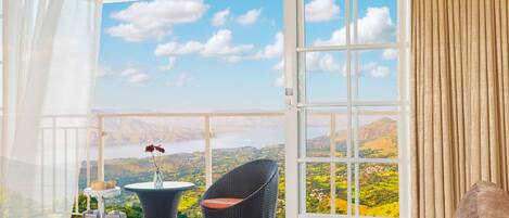 A balcony with a table set for tea, a wicker chair, and a sofa, overlooking a scenic landscape with mountains and a body of water.