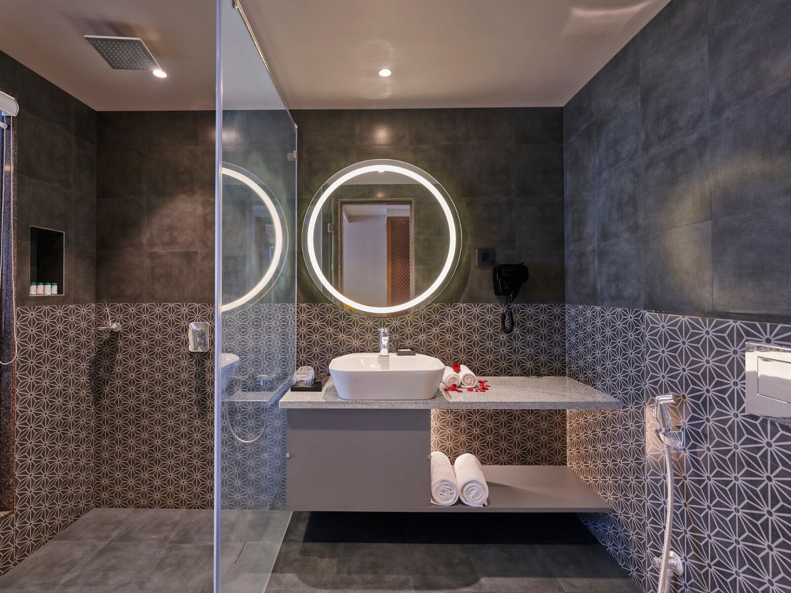 A modern bathroom with a glass shower enclosure, a round mirror with a light, a white sink, and a towel rack.