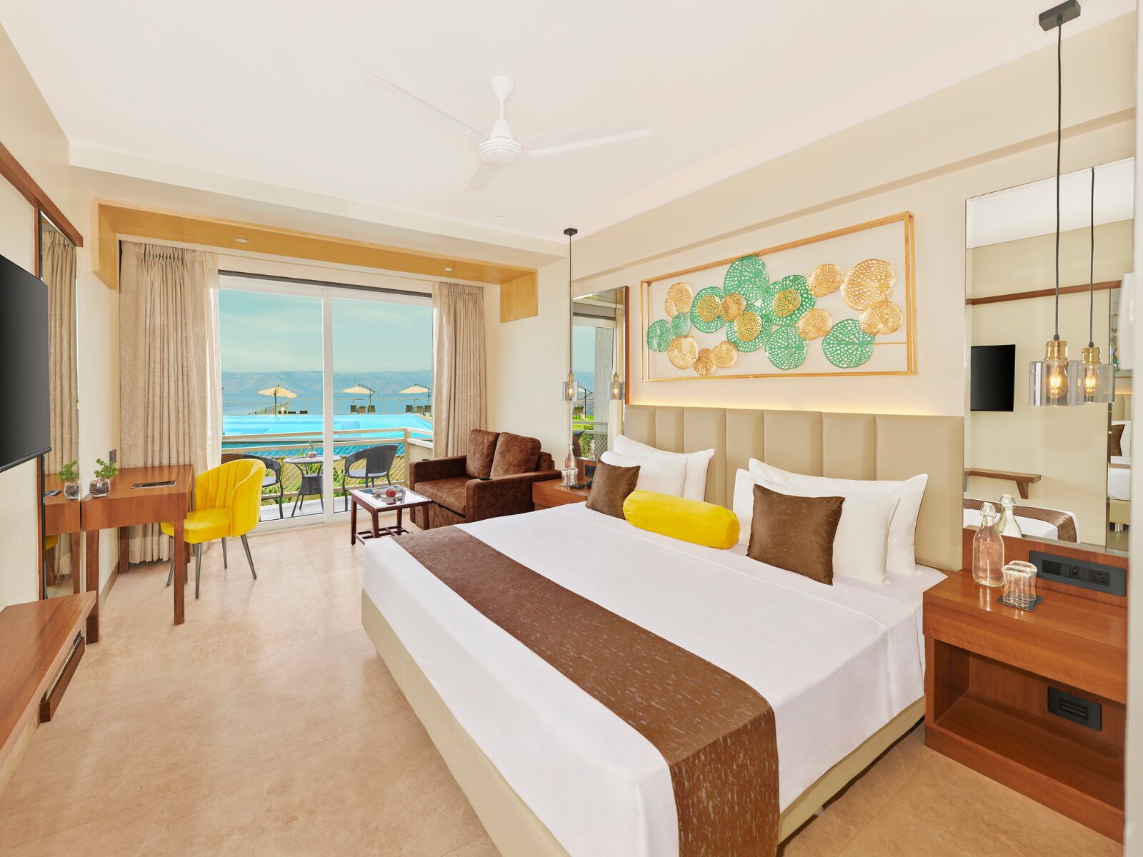 A hotel room with a large bed, a desk, a chair, a balcony with a view of the sea, and a ceiling fan.