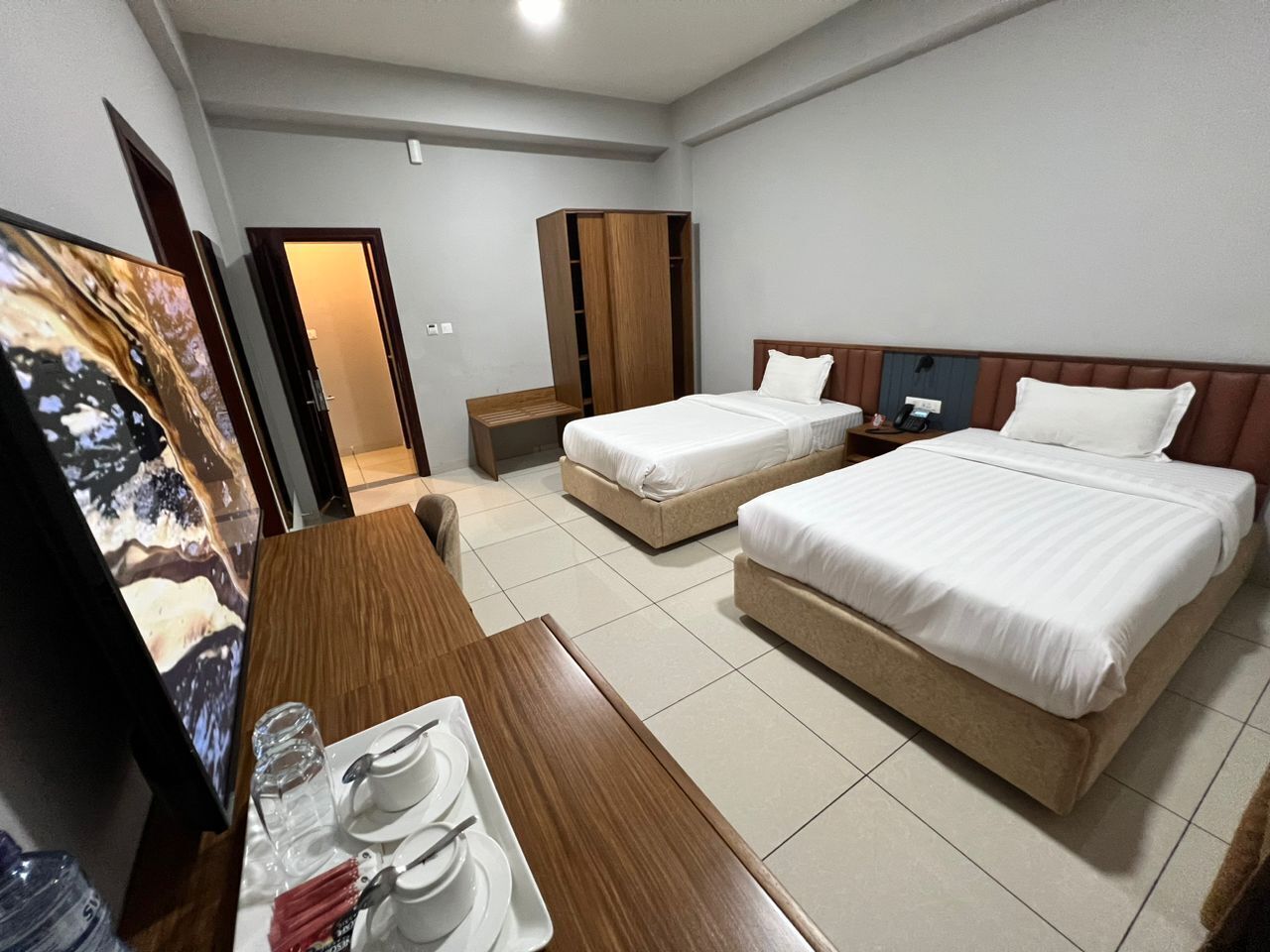 Une chambre d’hôtel avec deux lits, un bureau en bois sur lequel se trouve un plateau avec des tasses et des soucoupes blanches, et une armoire.