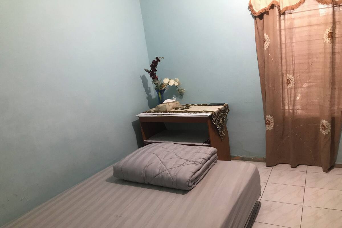 Un dormitorio pequeño y ordenado con una cama, una mesita de noche con un florero y una cortina.
