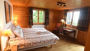Ein Zimmer einer Holzhütte mit einem Bett, zwei mit Vorhängen versehenen Fenstern, einem Schreibtisch und einem Stuhl.