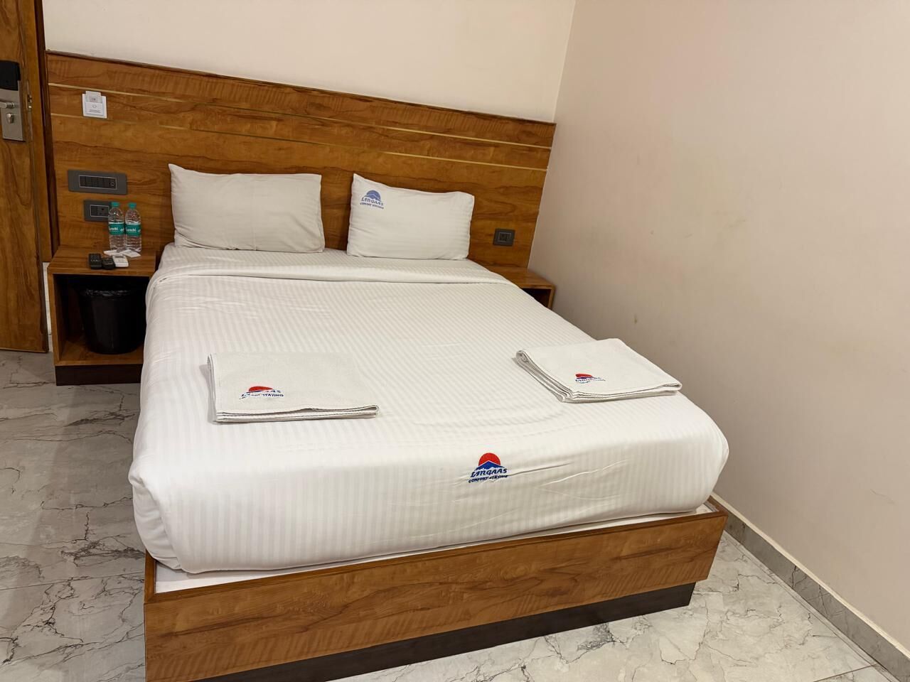 Una cama bien hecha con un colchón de marca, almohadas y toallas dobladas sobre un cabecero de madera en una habitación de hotel.