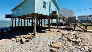 A house on stilts by the water with a wooden deck and a staircase leading to a second level.
