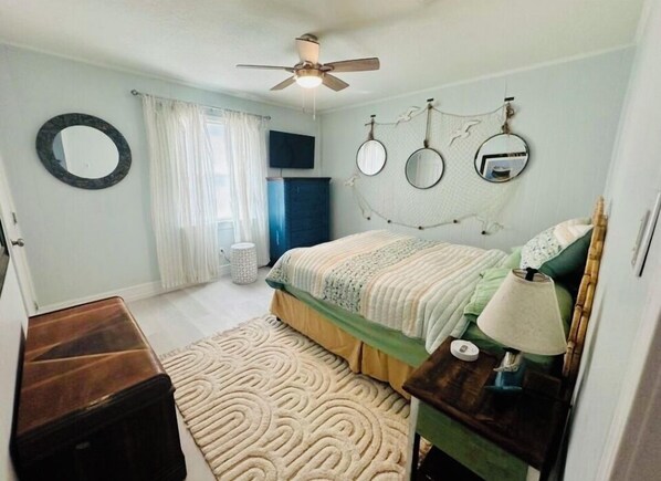 Un dormitorio con cama, una mesita de noche con lámpara, un espejo redondo en la pared, un ventilador de techo y una ventana con cortinas.