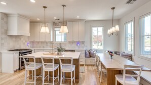 A modern kitchen with white cabinetry, a central island with bar stools, pendant lighting, and a view of the outdoors through large windows.