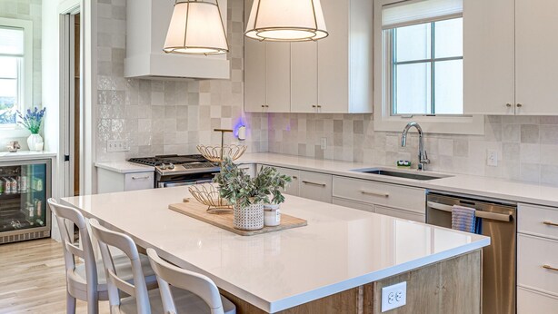 A modern kitchen with white cabinetry, a large island with a built-in sink, pendant lighting, and a view of the outdoors through a window.