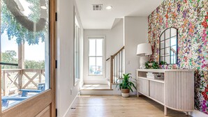 A hallway with wooden flooring, a staircase, a white console table with a mirror, and a colorful floral wall.