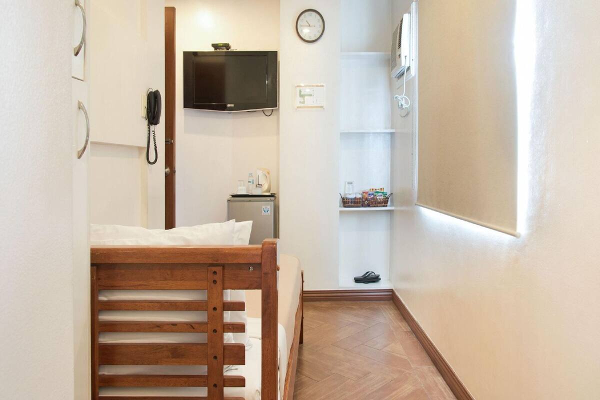 A small, narrow hotel room with a wooden bed, a television mounted on the wall, a small refrigerator, a shelf with toiletries, and a clock.