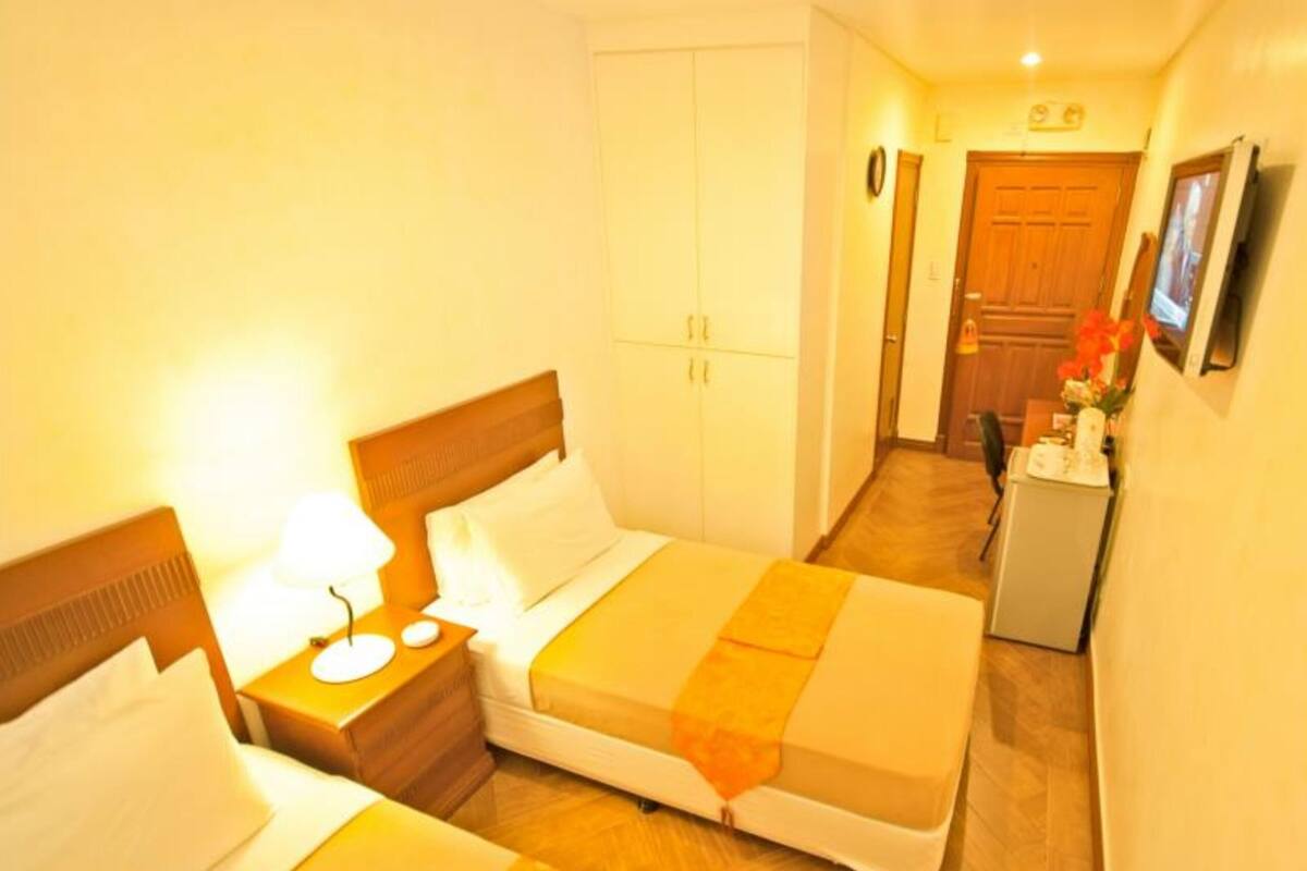 A hotel room with two beds, a nightstand with a lamp, a small table with a vase, and a TV on the wall.