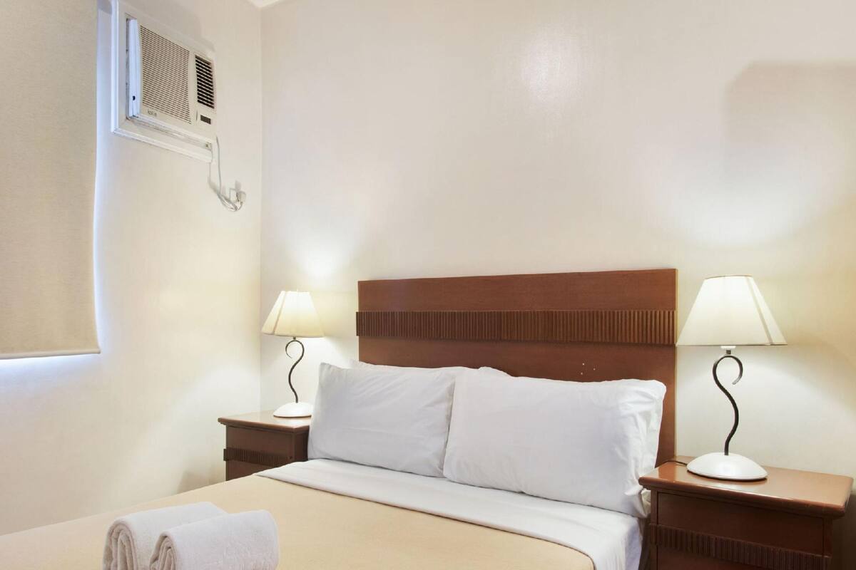 A hotel room with a bed, two bedside tables with lamps, a window with an air conditioning unit, and a towel on the bed.