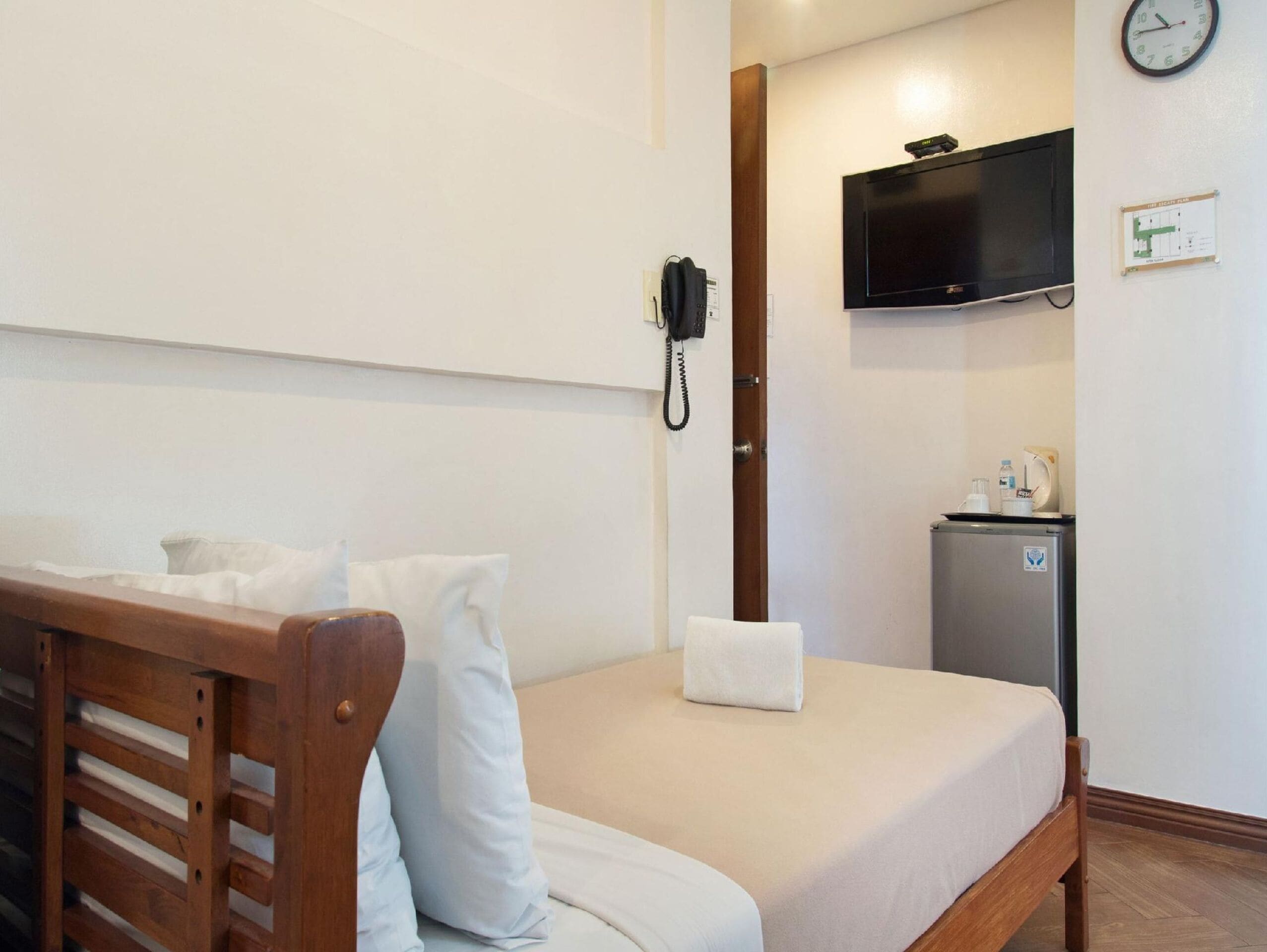 Habitación de hotel con cama de madera, ropa de cama blanca, un refrigerador pequeño, teléfono y televisor montado.