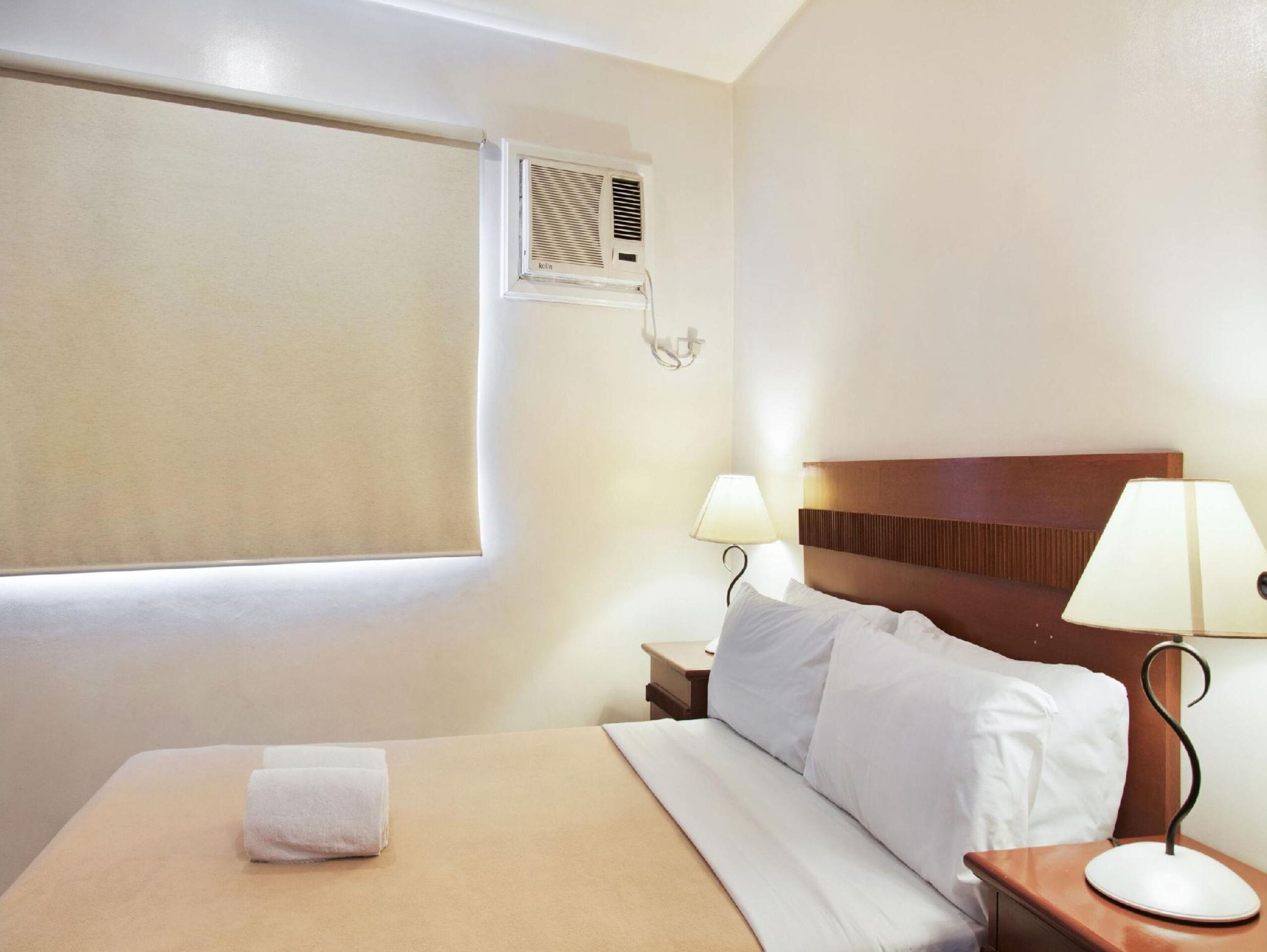 A hotel room with a bed, two bedside lamps, a nightstand, a window with blinds, and a wall-mounted air conditioning unit.
