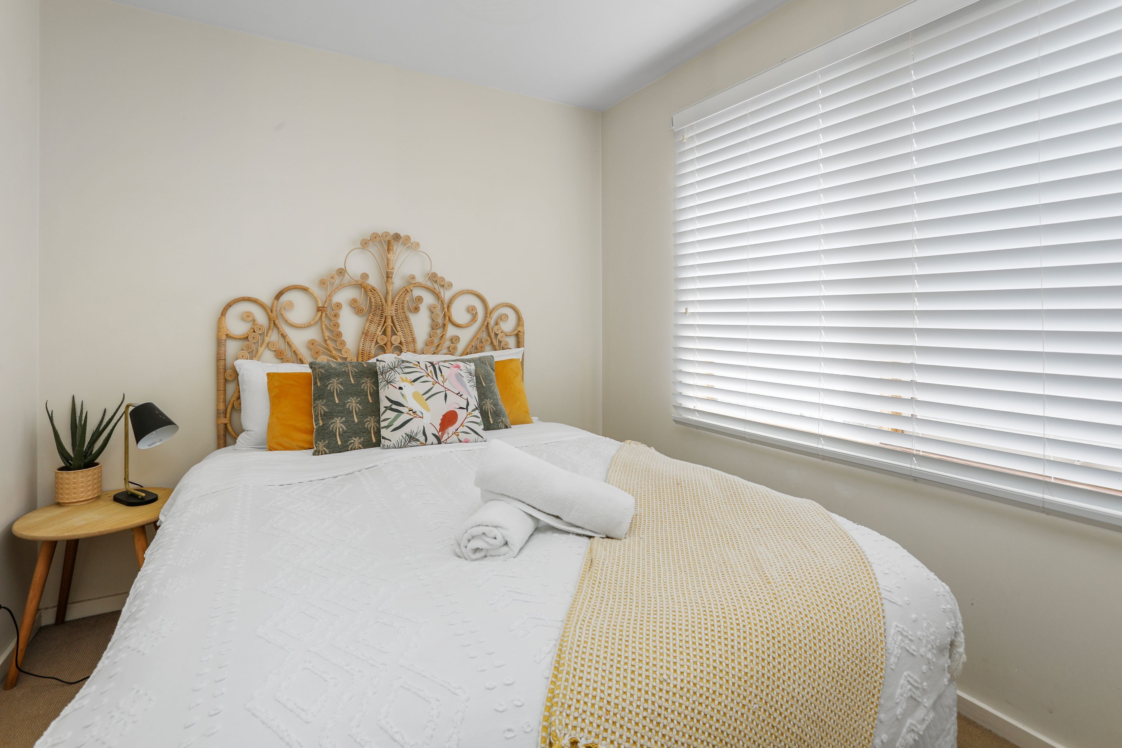A neatly made bed with a white duvet, a rolled white towel, and a yellow and white patterned throw blanket. There is a small bedside table with a lamp and a potted plant.