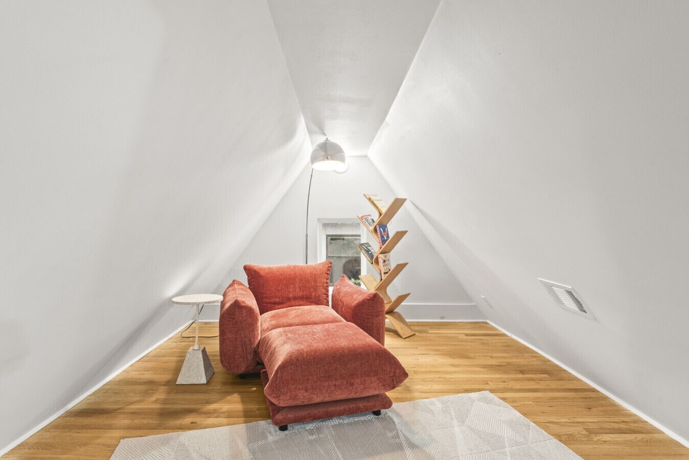 A modern, minimalist attic room with a red sofa, a small round table, and a wooden ladder bookshelf.