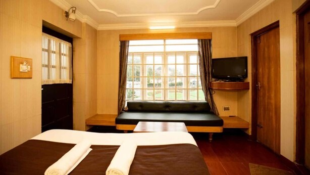 A hotel room with two beds, a large window with curtains, a television mounted on the wall, and a bench by the window. - Lazo Silver Lake (Ootacamund)