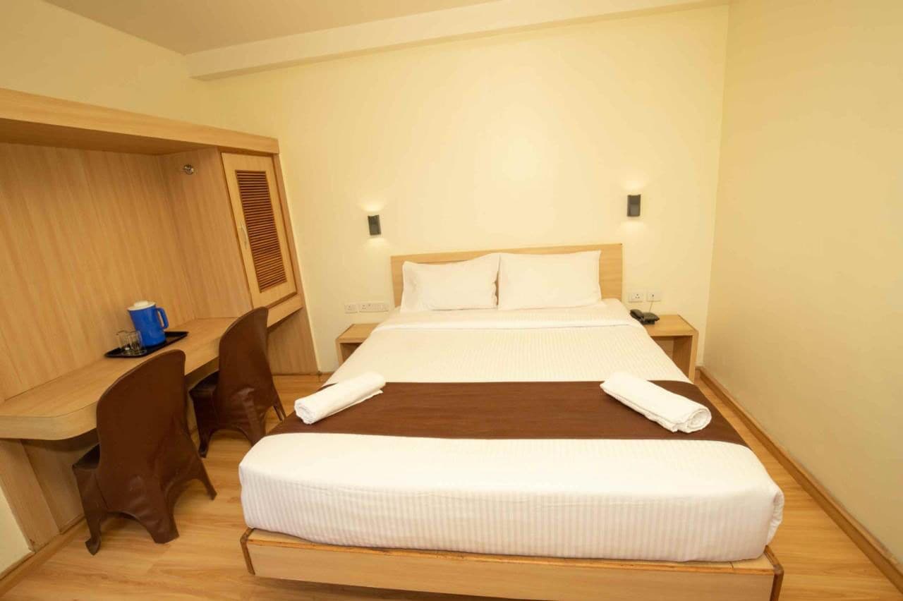 A hotel room with a bed, a desk, two chairs, and a small table with a blue cup and a glass.