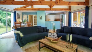 Un vaste espace de vie doté d’un haut plafond et de poutres apparentes, comprenant un canapé bleu foncé, une table basse en bois avec des bougies et de grandes fenêtres offrant une vue sur l’extérieur.
