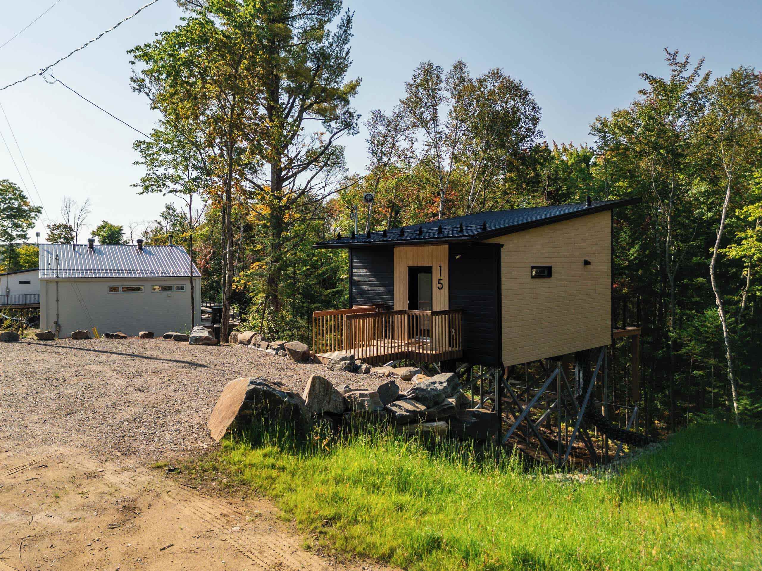 A cabin with a wooden deck and a small porch, surrounded by trees and a gravel path.