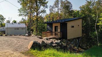 A cabin with a wooden deck and a small porch, surrounded by trees and a gravel path.