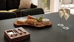 A table with a cheese board, grapes, and two glasses of white wine, set in a room with a view of trees through large windows.